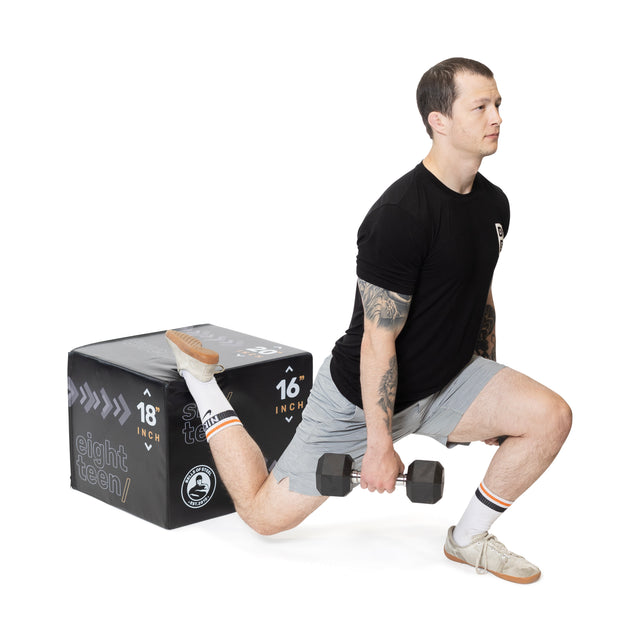 A person executes a Bulgarian split squat, holding dumbbells in each hand with their back foot elevated on an Bells of Steel Foam Soft Plyo Box (16 x 18 x 20). Wearing a black t-shirt, gray shorts, and white socks and shoes, they demonstrate the versatility of their home gym setup.