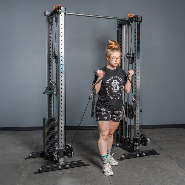 A woman in glasses, a black T-shirt, patterned shorts, and sneakers performs bicep curls with both hands using the Bells of Steel Cable Tower Squat Stands in a gym with a gray wall.