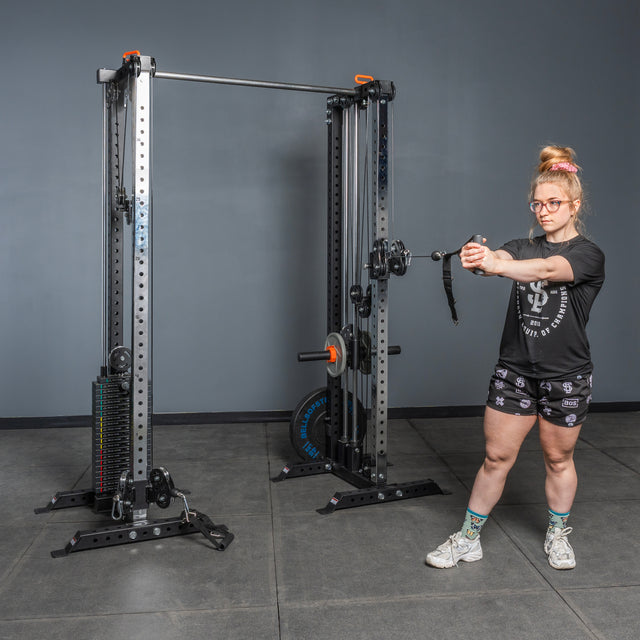 A woman in athletic wear uses the Bells of Steel Cable Tower Squat Stands in a gym, pulling the handle with both hands while standing sideways.