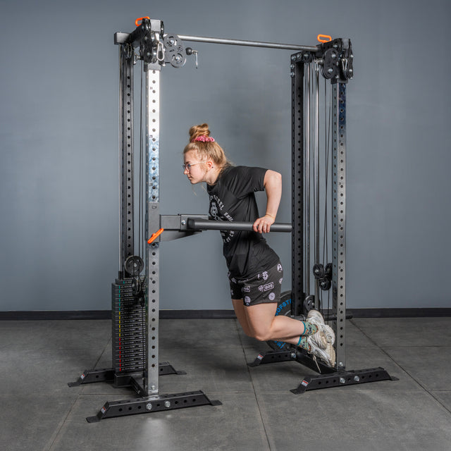 A person with light hair in a bun, glasses, black t-shirt, and shorts performs a dip exercise on the Bells of Steel Cable Tower Squat Stands in a gym setting.