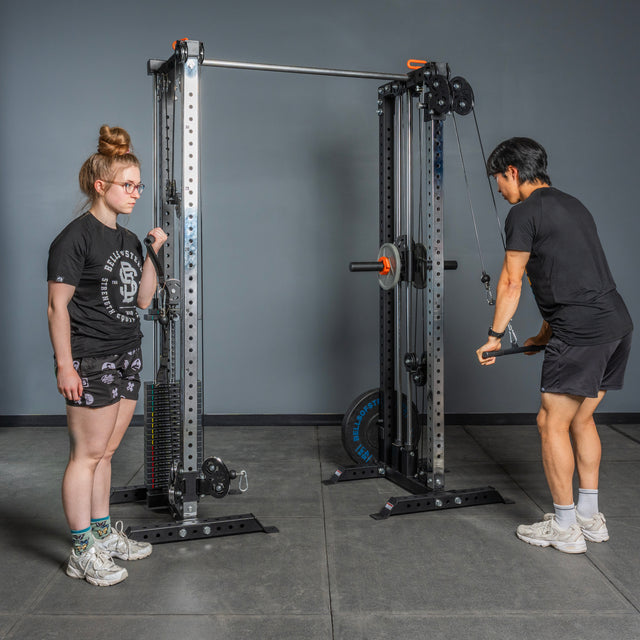 Two people use the Bells of Steel Cable Tower Squat Stands in the gym. One stands facing forward, arms at her sides, while the other pulls a cable down with both hands. Both wear athletic clothes and sneakers; backdrop is a gray wall.