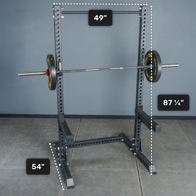 The Bells of Steel Manticore Squat Stand - Prebuilt (3" x 3", 1" Holes) features sturdy black metal construction with robust 3x3 uprights and a barbell loaded with weights. Measuring 87¼" in height, 49" in width, and 54" in depth, this squat stand is set on a tiled floor against a gray wall—making it an ideal addition to any home gym.