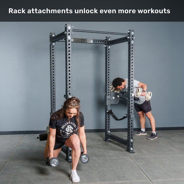 Two people use the Bells of Steel Manticore Four Post Power Rack – Prebuilt (3" x 3", 1" Holes) with attachments; one does lunges holding dumbbells, while the other performs a bent-over row with a barbell. Text above reads: “Rack attachments unlock even more workouts—perfect for building your home gym.”.