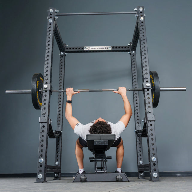 A person lies on a bench and grips a barbell with weights, preparing to bench press inside the Manticore Four Post Power Rack - Prebuilt (3" x 3", 1" Holes) by Bells of Steel in a gym setting.