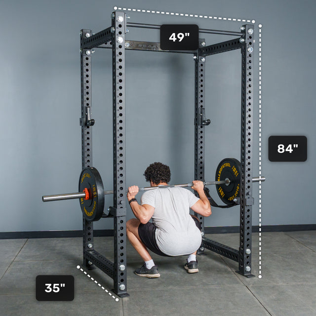 A person performs a squat inside the Bells of Steel Manticore Four Post Power Rack - Prebuilt (3" x 3", 1" Holes), shown here in black with dimensions clearly labeled: 49 inches wide, 35 inches deep, and 84 inches tall. With weight plates loaded on the barbell, this modular rack is an excellent addition to any home gym.