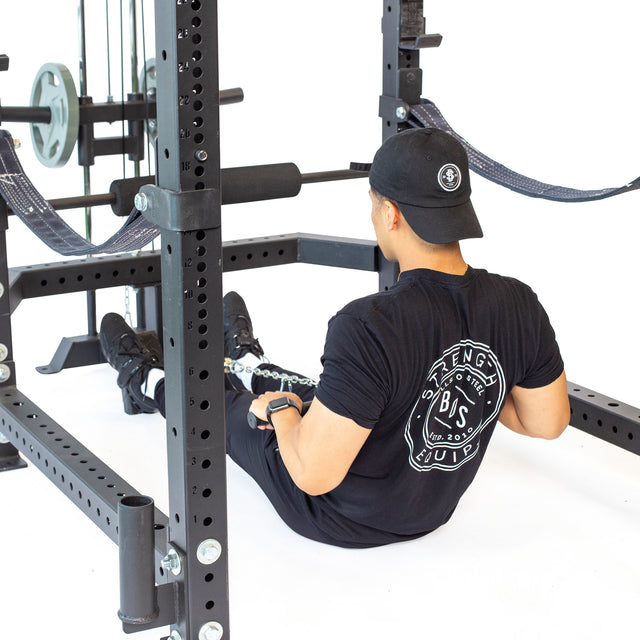 Wearing black workout clothes and a cap, a person sits on the gym floor using the Bells of Steel Lat Pulldown & Low Row Rack Attachment—Hydra & Manticore for seated rows. Olympic Weight Plates and other equipment are visible nearby.