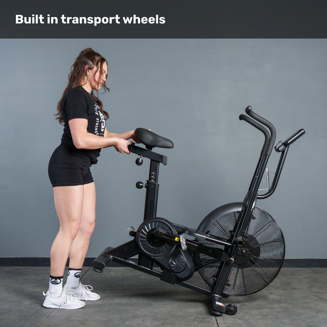 A woman in workout gear moves the Bells of Steel Residential Air Bike with Wind Guard using its built-in transport wheels. The text above reads, "Built in transport wheels"—ideal for any home gym.