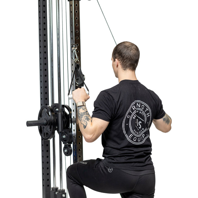 A tattooed person in a black "Strength Equipment" shirt uses Bells of Steel Adjustable Length Aluminum Handles on a cable machine for a lat pulldown, kneeling with a white background that highlights these premium gym accessories.