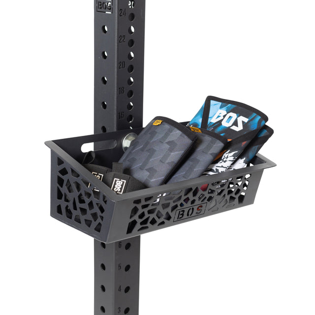 The Bells of Steel Rack Attached Storage Basket mounts to a gym rack post, organizing accessories like wrist wraps and a water bottle. Its cutout geometric pattern and BOS branding make it perfect for efficient gym storage.