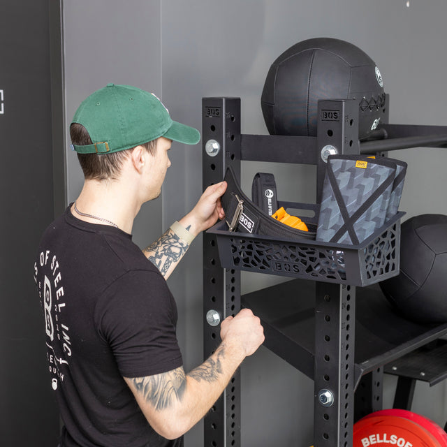 A man in a green cap and black t-shirt arranges fitness gear on a Bells of Steel Rack Attached Storage Basket, which is part of a gym storage rack also holding medicine balls.