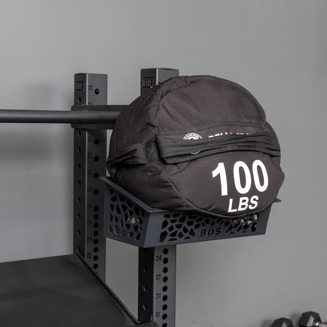A black "100 LBS" weighted sandbag rests on a Bells of Steel Rack Attached Storage Basket, which is part of a metal gym storage system against a gray wall.