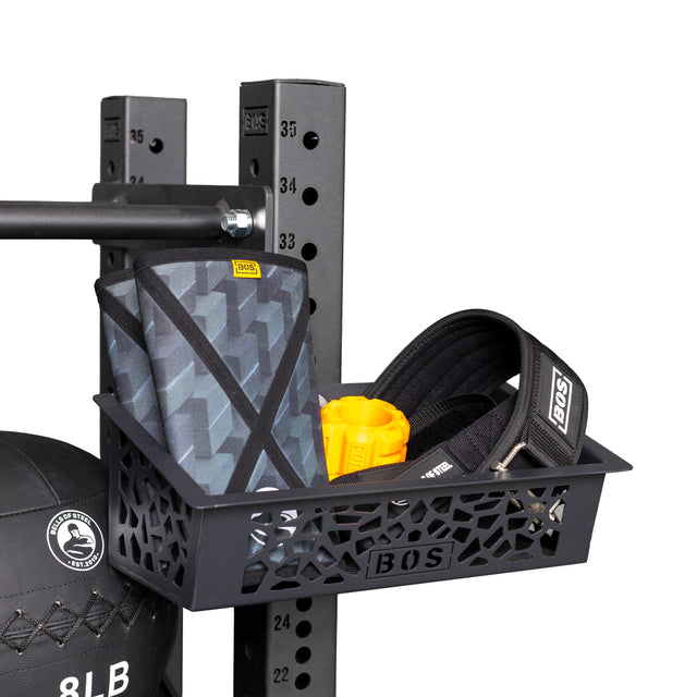The Bells of Steel Rack Attached Storage Basket keeps a black exercise belt, patterned grey knee sleeve, yellow handle, and more organized. A black medicine ball sits partially visible below the basket.