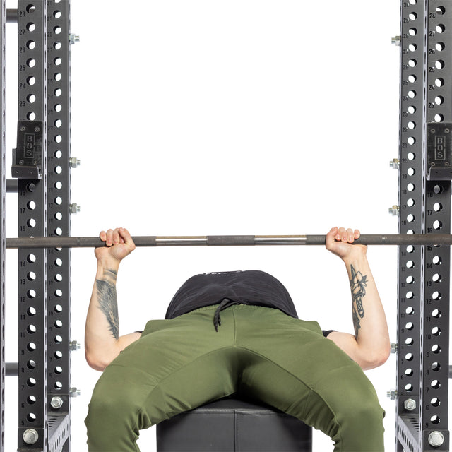 Wearing green pants, a person lies on a bench in a power rack with black metal supports and Bells of Steel Standard J-Cups, gripping a barbell above their chest as they get ready to bench press.