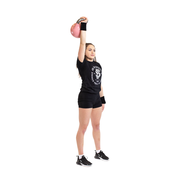 A person stands on a white background holding a pink kettlebell overhead, wearing Bells of Steel’s BellGuard Kettlebell Wrist Guards (Pair) along with a black t-shirt, black shorts, and black athletic shoes. Perfect for kettlebell lifters.