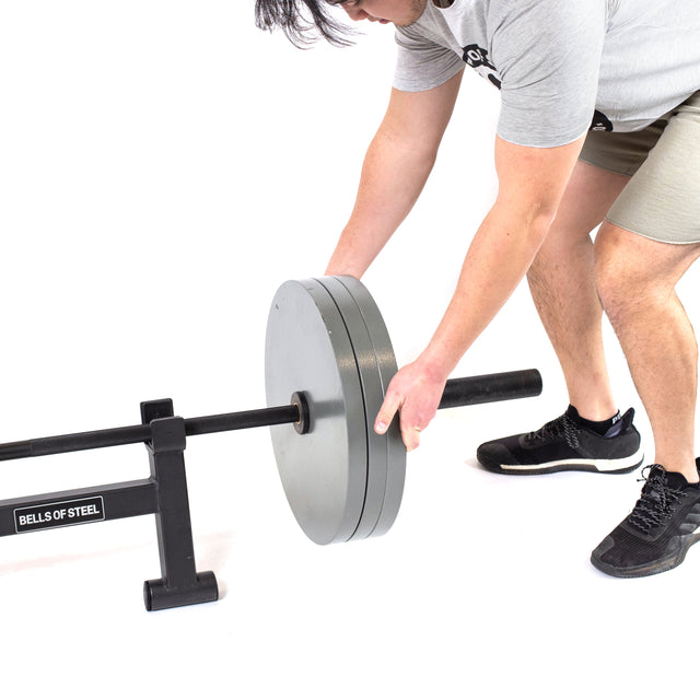A person in athletic wear loads large weight plates onto a Bells of Steel Powerlifting Bar on a rack, set against a plain white background.