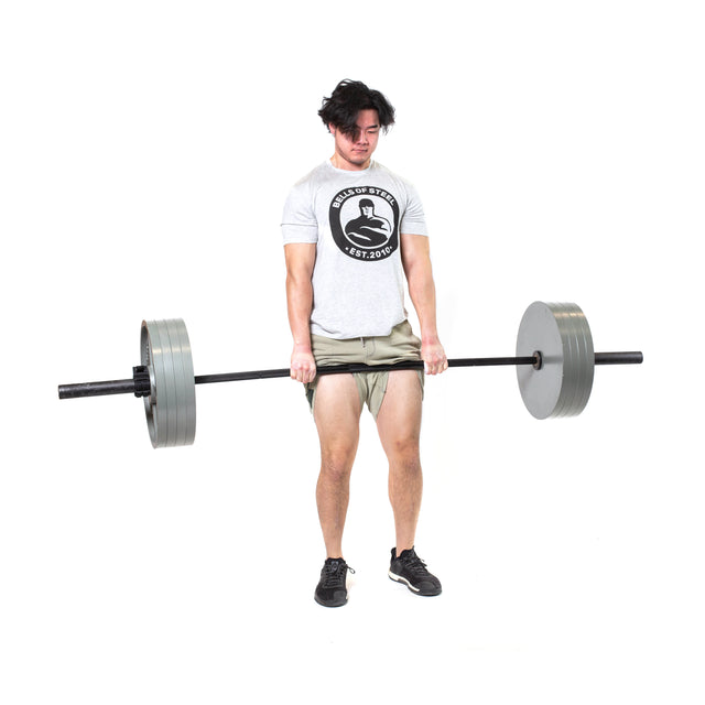 A man in a gray t-shirt and shorts lifts the Bells of Steel Powerlifting Bar, built to IPF specs. He stands on a white background, gripping the bar with both hands and displaying effort on his face.