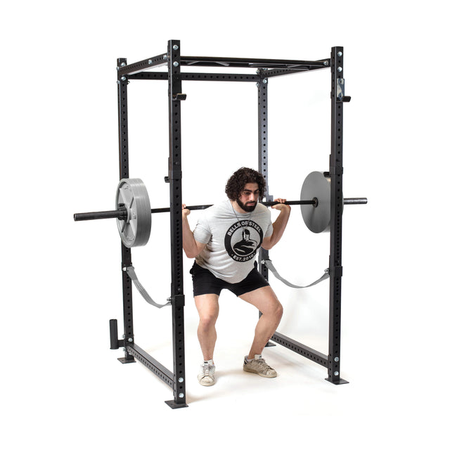 A man with curly hair and a beard performs a barbell squat inside a rack, using the Bells of Steel Powerlifting Bar. He wears a white T-shirt, black shorts, and sneakers. The rack features safety straps and large weight plates on each side.