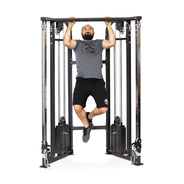 A bearded man in a gray t-shirt, black shorts, and black shoes does a pull-up on the Bells of Steel Functional Trainer - Weight Stack, featuring dual weight stacks and cables for versatile exercises.