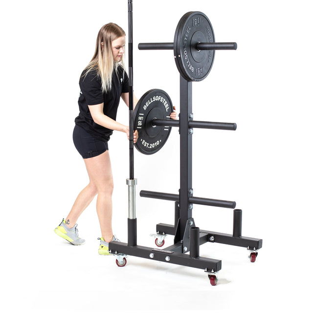 A person places a weight plate onto a horizontal storage peg of the Bells of Steel Bumper Plate Weight Tree And Bar Holder, which holds multiple plates and a vertical barbell, set against a white background.