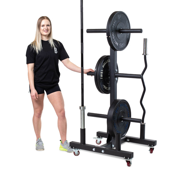A smiling woman in athletic wear stands next to the Bells of Steel Bumper Plate Weight Tree and Bar Holder, which stores weight plates, a barbell, and a hex bar on wheels against a white background.