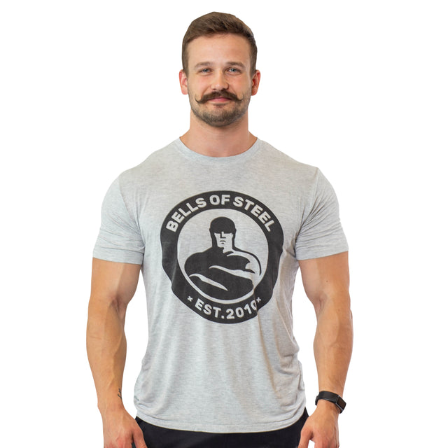 A mustached man smiles against a white background, wearing a light gray Bells of Steel Bamboo T-Shirt featuring the "Bells of Steel Est. 2010" logo.