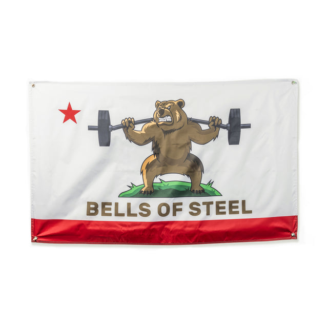 The Bells of Steel Flags feature a cartoon bear on grass lifting a barbell, perfect for gym decor. A red star is in the top left corner, with "BELLS OF STEEL" boldly printed at the bottom over a red stripe.