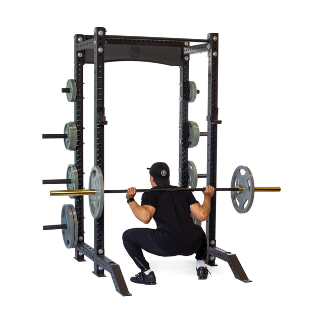 A person in black workout clothes and a cap performs a barbell squat viewed from behind using the Bells of Steel Hydra Collegiate Power Rack - Prebuilt (3" x 3", ⅝" Holes), highlighting essential modular gym equipment for any home gym, set against a white background.