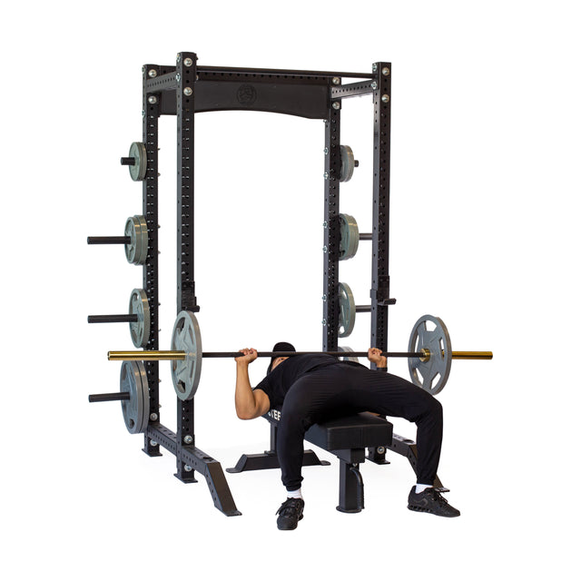 Wearing black athletic wear, a person bench presses with a barbell inside the Bells of Steel Hydra Collegiate Power Rack (Prebuilt, 3" x 3", ⅝" Holes), with weight plates stored on side pegs against a plain white background.