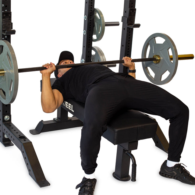 Wearing black athletic wear, a person prepares to lift a loaded barbell on a bench inside the Bells of Steel Hydra Collegiate Power Rack (Prebuilt, 3" x 3", ⅝" holes)—an ideal addition to any home gym.