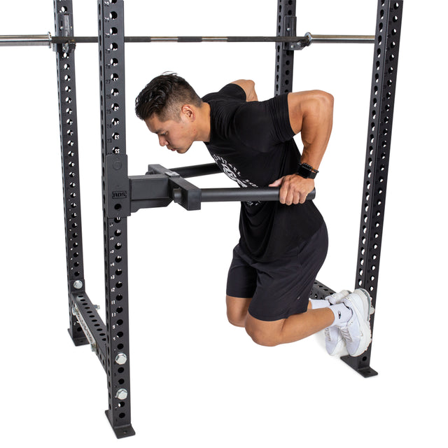 A man in a black T-shirt and shorts uses a Bells of Steel Y Dip Bar Rack Attachment connected to a power rack for dip exercises. In the gym, he bends his knees and keeps his feet off the ground for an intense workout session.