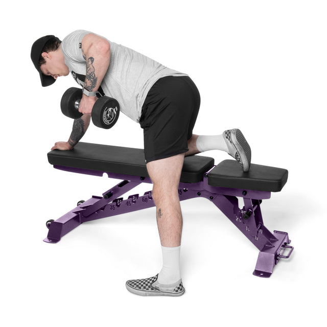 Wearing a gray shirt, black shorts, and a black cap with patterned slip-on shoes, a person performs a one-arm dumbbell row on the Buzz-Saw Heavy-Duty Adjustable Bench by Bells of Steel, using the purple bench for support.