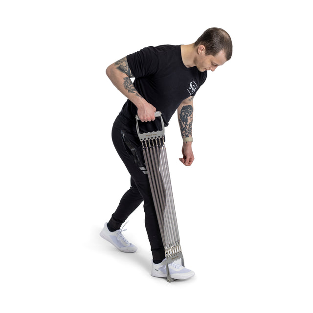 A tattooed individual in a black t-shirt and pants uses the Bells of Steel Chest Expander, pulling it downwards with one hand on a white background, embodying classic bodybuilding style.