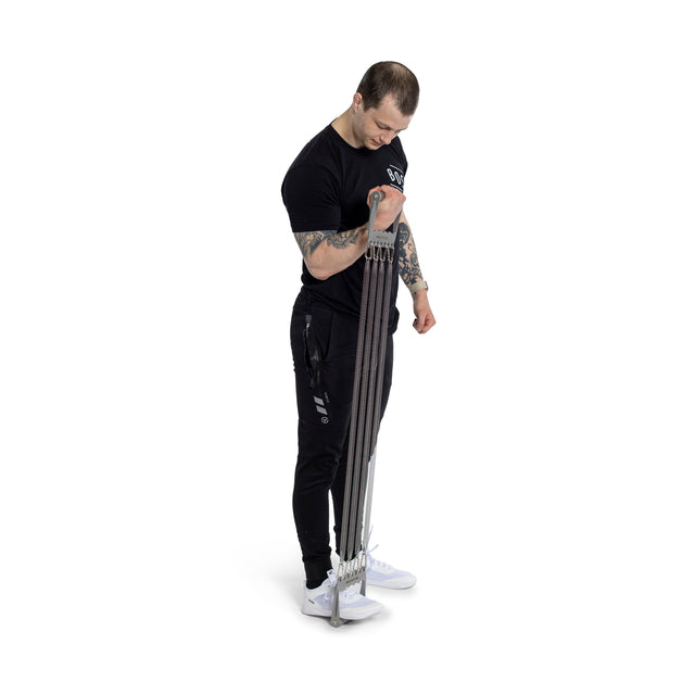 A person wearing a black shirt and pants uses the Bells of Steel Chest Expander for bicep curls, showcasing adjustable resistance. Visible arm tattoos and white sneakers create a focused strength-training scene against a plain background.