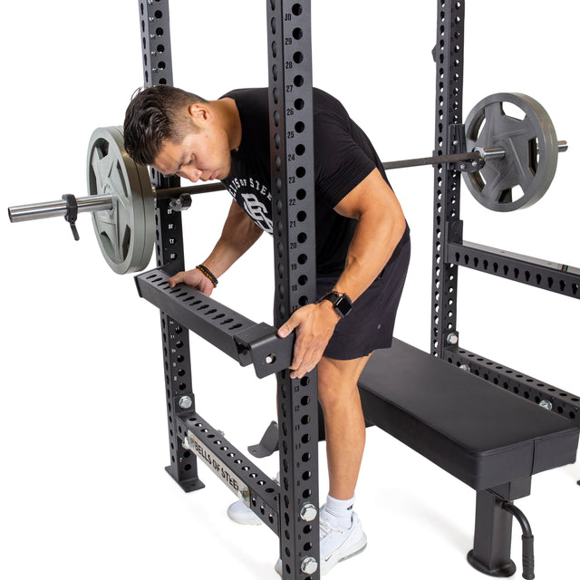 A man in black athletic wear and white sneakers adjusts Bells of Steel Flip-Down Safeties on a power rack next to a loaded barbell and bench, prioritizing safe training before his workout.
