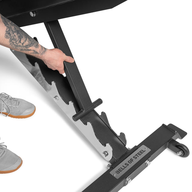 A person adjusts a Bells of Steel Flat/Incline/Decline Weight Bench with a silver frame, expertly altering its angle for optimal muscle targeting. A tattooed arm and grey sneakers are partially visible beside this versatile commercial exercise station.