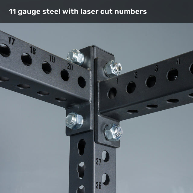 Close-up of intersecting black steel beams with holes and precision laser-cut numbers, secured by large bolts. Text above reads, "11 gauge steel with laser cut numbers." The robust construction of the Bells of Steel Manticore Flat Foot Power Rack (3" x 3", 1" Holes) makes it an ideal centerpiece for any home gym.