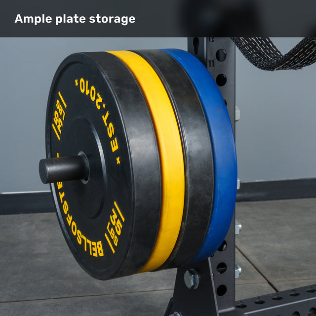 Black, yellow, and blue weight plates are neatly stacked on a horizontal rack attached to the Bells of Steel Manticore Half Rack Builder (3" x 3", 1" Holes) with sturdy 3x3 uprights, set on a gray floor, with the text "Ample plate storage" above.
