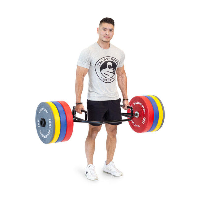 A man in a gray Bells of Steel shirt and black shorts lifts the Bells of Steel Open Trap Bar / Hex Bar with rotating sleeves and colorful weight plates, standing on a white background.