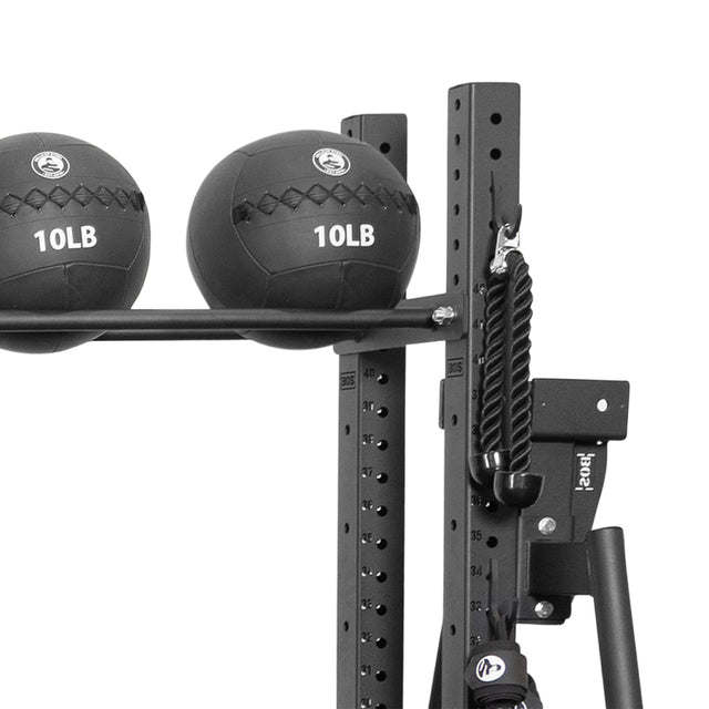 Two black 10-pound medicine balls rest on a Bells of Steel Utility Hook for cable attachments, all beside a rope on a black metal gym frame with numbered holes—a perfect storage solution.
