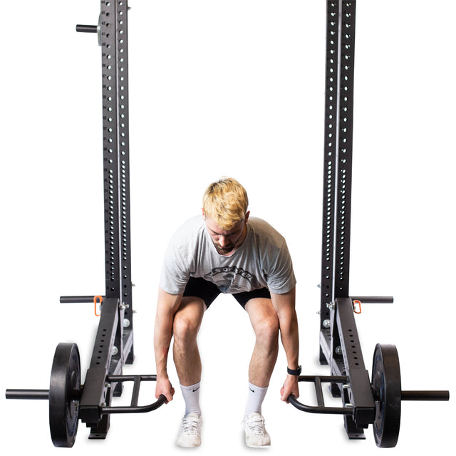 A person in athletic wear prepares to use the Bells of Steel Lever Arms Rack Attachment inside a squat rack, viewed from the front against a white background.