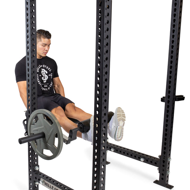 A person in a black t-shirt and shorts uses the Bells of Steel Leg Curl / Leg Extension Rack Attachment, performing a leg extension with their legs extended and back supported.