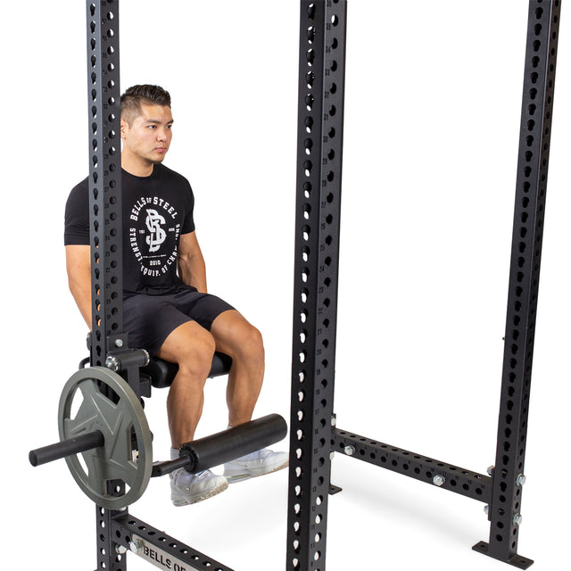 A man in athletic wear sits on a bench inside a squat rack, holding the seat as weight plates are loaded on the Bells of Steel Leg Curl / Leg Extension Rack Attachment in front of him.