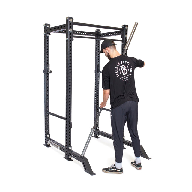 Wearing a black Bells of Steel shirt and cap, someone places a barbell inside a power rack with Bells of Steel Front Foot Stabilizers and multiple adjustment holes, getting ready for their workout.