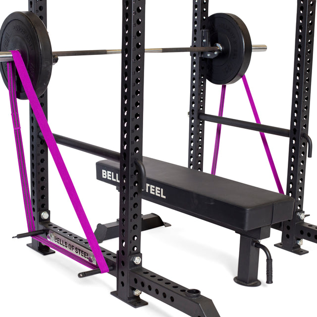 A Bells of Steel bench press setup with a barbell and weights inside a power rack, using Standard Band Pegs to secure two purple resistance bands from the base to the barbell. The bench is branded "BELLS OF STEEL.