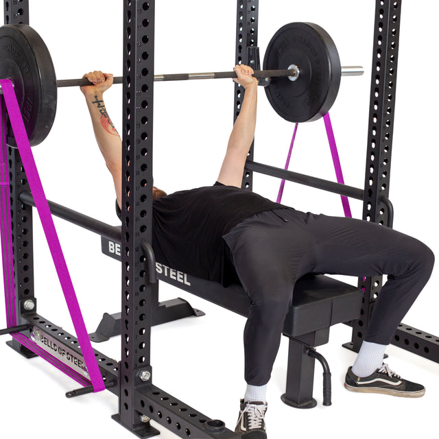 Wearing black workout gear and sneakers, a person does a barbell bench press in a power rack using Bells of Steel Standard Band Pegs for resistance, with safety straps on both sides for protection.