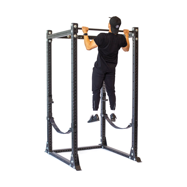 Wearing black athletic wear and a cap, a person performs a pull-up on the Bells of Steel Hydra Flat Foot Power Rack Builder (3" x 3", ⅝" Holes), demonstrating its sturdy design against a white background—ideal for any home gym.