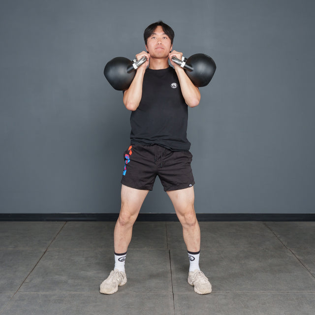 A person in black workout gear stands on a gym floor holding two Bells of Steel Competition Kettlebells at shoulder height, preparing to exercise.