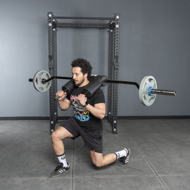 A man in athletic wear uses the Bells of Steel Safety Squat Bar – The SS4 for lunges, holding it across his shoulders in front of a squat rack, with his right knee bent forward and left leg extended behind.