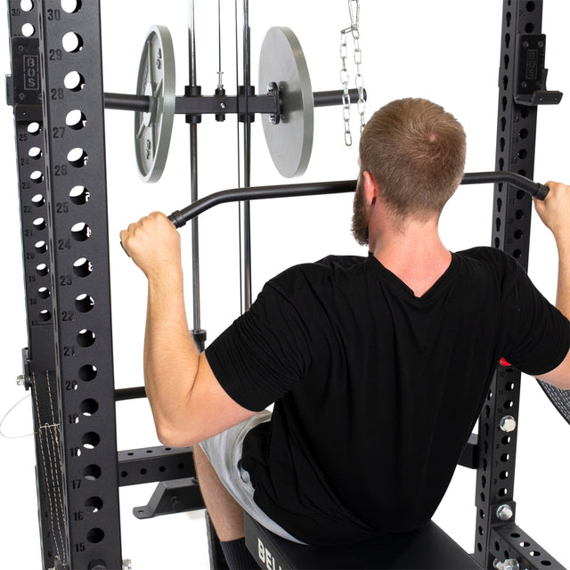 A bearded man with short hair, wearing a black shirt and gray shorts, uses the Bells of Steel Lat Pulldown & Low Row Rack Attachment – Hydra & Manticore with Olympic Weight Plates in a power rack for cable exercises.