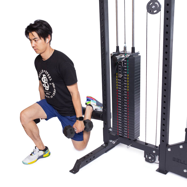 A man in athletic wear uses the Bells of Steel Lat Pulldown Low Row Machine for support while performing a Bulgarian split squat with dumbbells, demonstrating this equipment’s versatility for both cable and strength exercises at home.